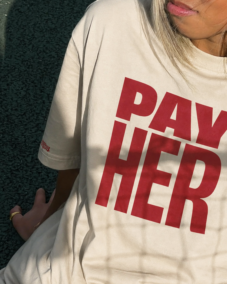 A woman wearing a cream-colored oversized t-shirt with the bold red text 'PAY HER' printed on the front, set against a dark textured background with a prominent shadow.