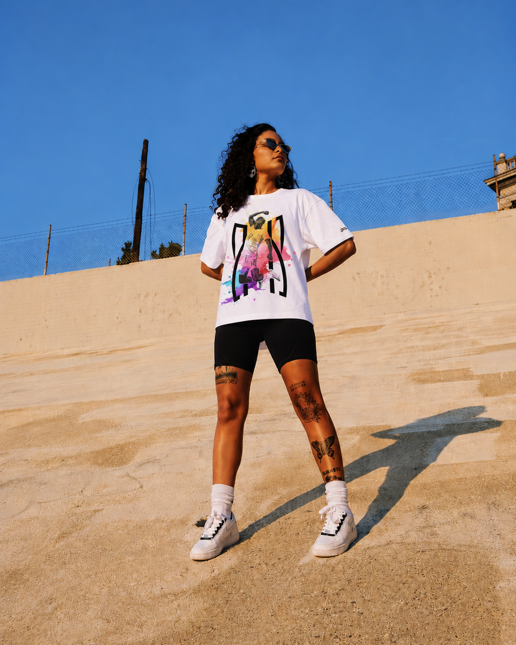 Person wearing a white t-shirt with a colorful graphic design, black shorts, and white sneakers on a sandy surface with a clear blue sky.
