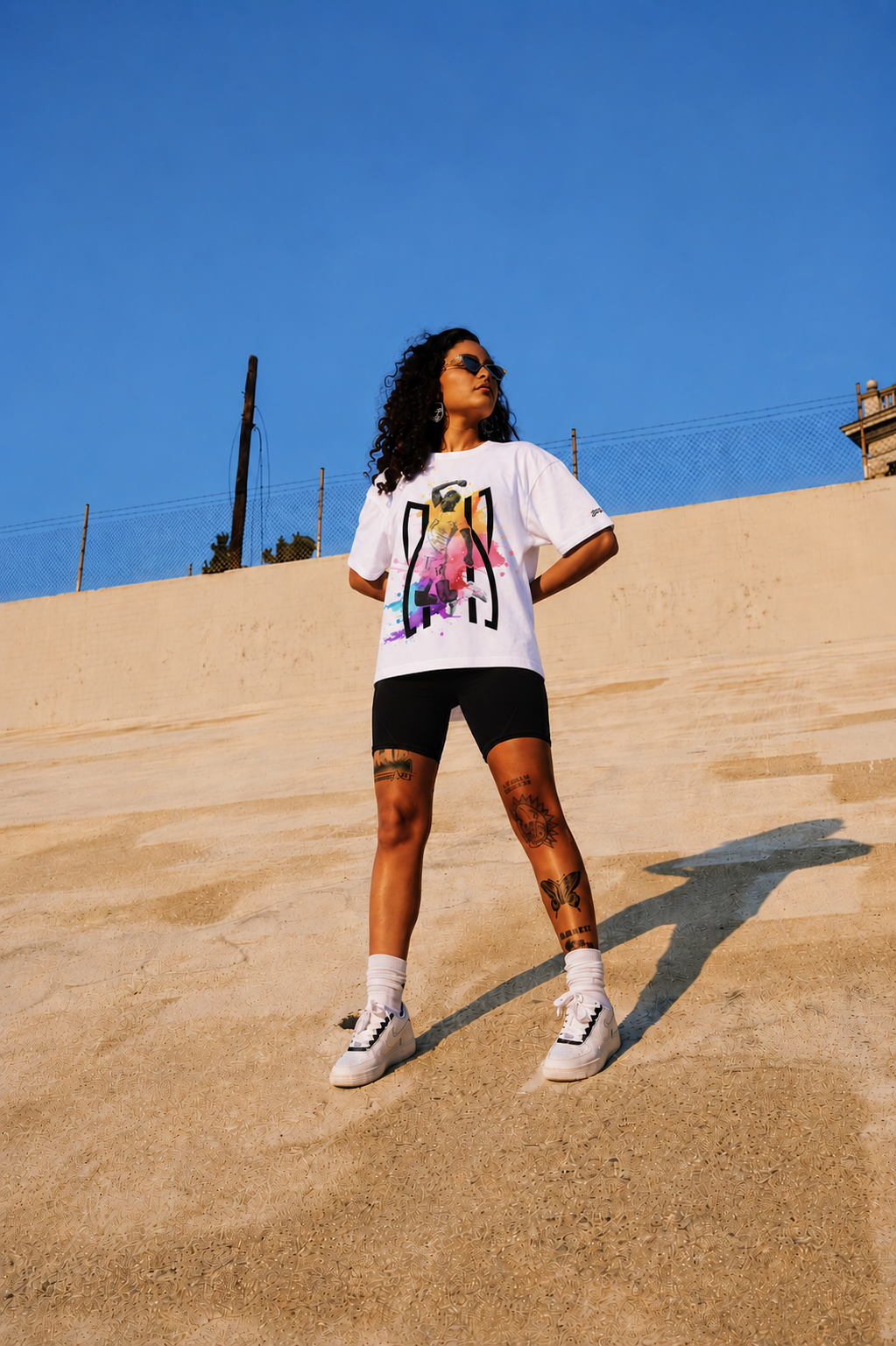 Person wearing a white t-shirt with a colorful graphic design, black shorts, and white sneakers on a sandy surface with a clear blue sky.