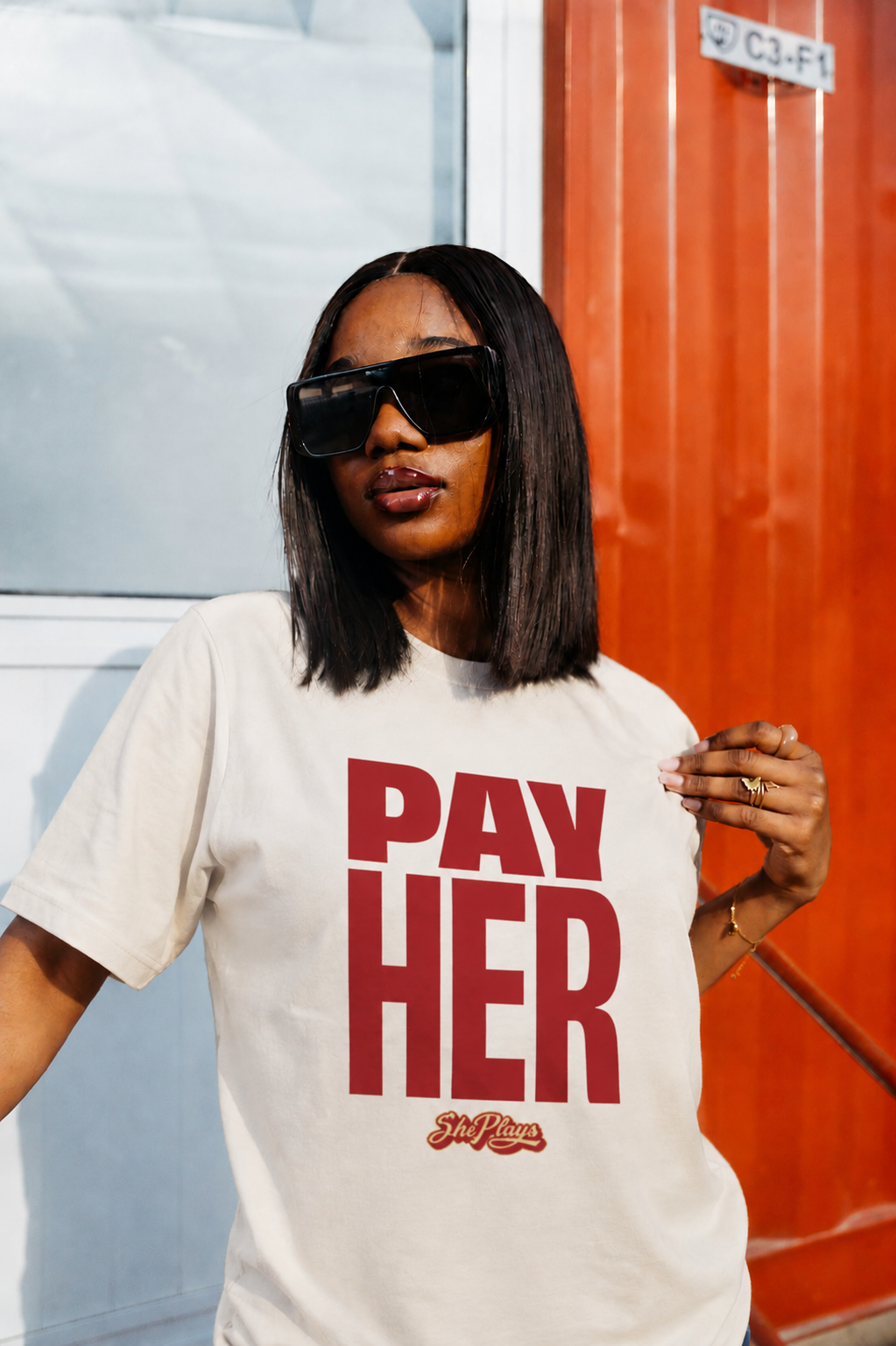 Person wearing a white t-shirt with 'PAY HER' in red text, standing in front of a red and gray wall.