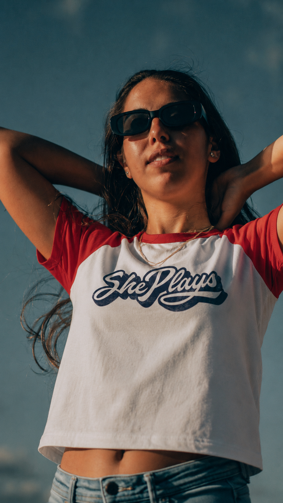 Woman wearing a red and white t-shirt with 'ShePlays' logo against a blue sky.